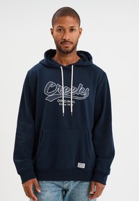 Sudadera azul marino con un bolsillo tipo canguro en la parte frontal, cordones blancos y la frase "Creeks Original Desde 1974" impresa en blanco en el pecho.