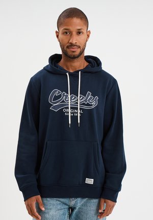 Marineblaues Hoodie mit einer vorderen Kängurutasche, weißen Zugbändern und dem Aufdruck "Creeks Original Since 1974" in Weiß über der Brust.