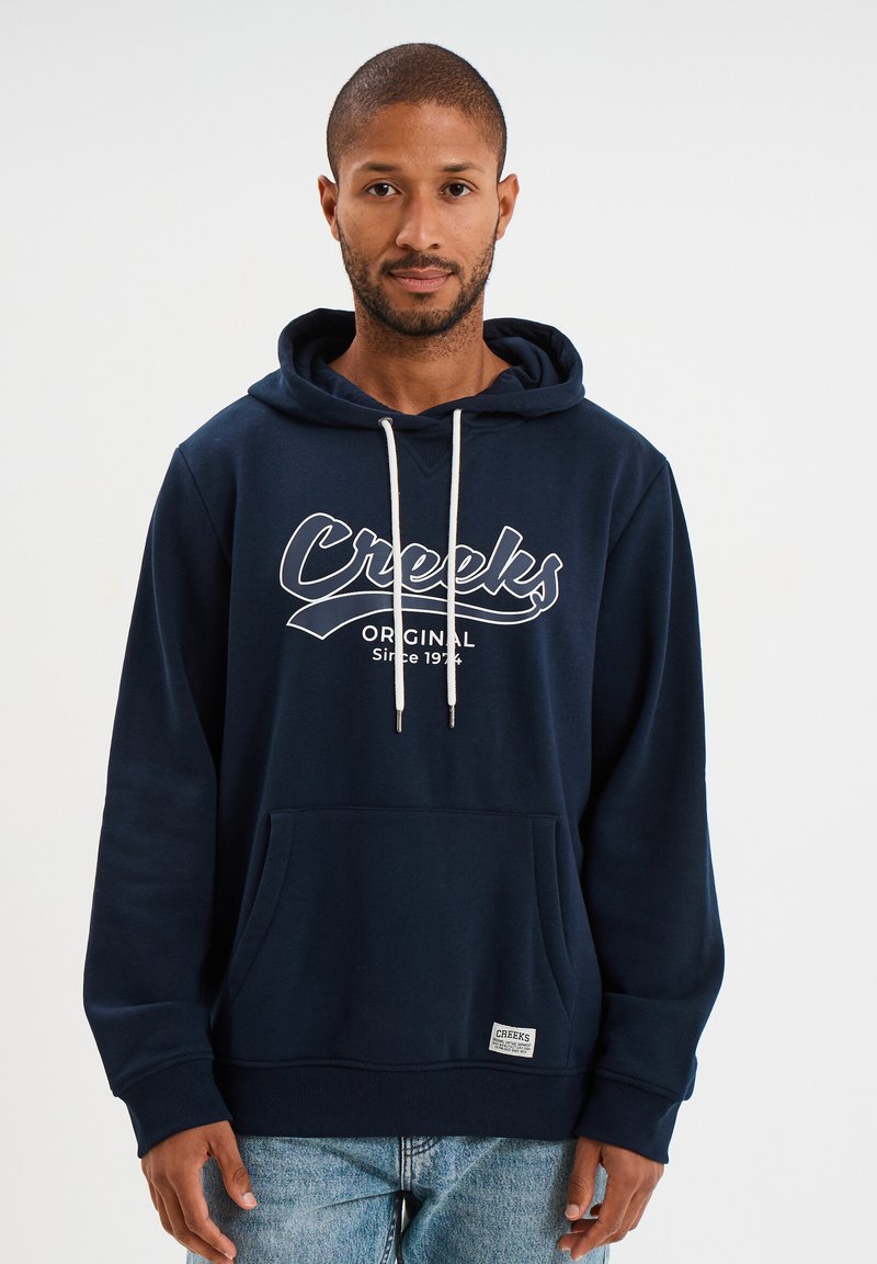 Sudadera azul marino con un bolsillo tipo canguro en la parte frontal, cordones blancos y la frase "Creeks Original Desde 1974" impresa en blanco en el pecho.