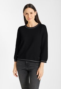 Vrouw met recht, donker haar, die een zwarte trui met grijze afwerking en donkergrijze jeans draagt, staande tegen een witte achtergrond.