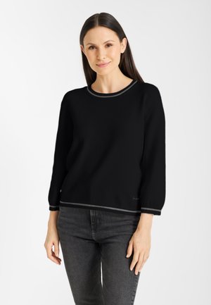Vrouw met recht, donker haar, die een zwarte trui met grijze afwerking en donkergrijze jeans draagt, staande tegen een witte achtergrond.