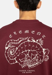 Bordeaux sweatshirt met een witte graphic van een krokodil en de tekst "CREEP, CRAWL, OR ROLL" gedrukt op de achterkant. Zacht, textuurstof.