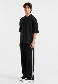 Sort polo-shirt med korte ærmer og krave, parret med løse sorte sweatpants med en lys mønstret sidestreg og hvide sneakers.
