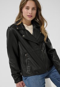 Melnā ādas jaka ar asimetrisku rāvējslēdzēju, pogām ar snap mehānismu un divām rāvējslēdzēju kabatām. Stilizēta ar krēma krāsas džemperi zem tās.