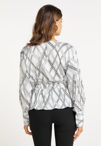 Weiße Bluse mit langen Ärmeln und schwarzem Kettenmuster, tailliert und mit elastischer Raffung am Saum. Kombiniert mit schwarzen, taillierten Hosen.