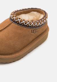 Bruna mockasiner i mocka med en vadderad insida, dekorativ flätad mönster vid kragen och en strukturerad gummisula. UGG-logotyp synlig.