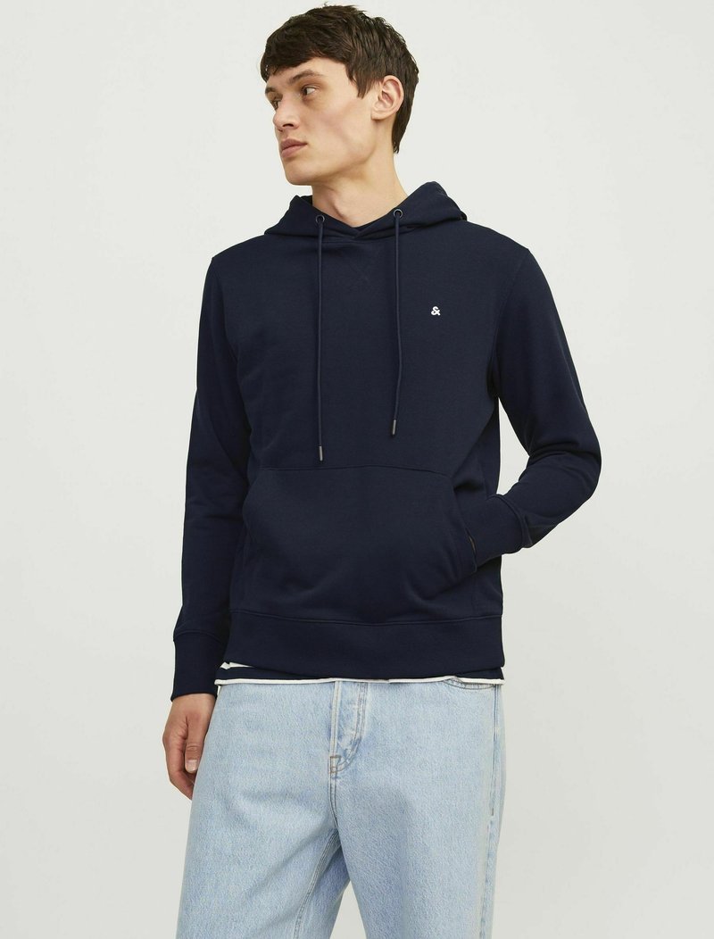 Marineblaue Kapuzenjacke aus weichem Stoff, mit einer Vorderaschtasche, Kordelzügen und einem kleinen Logo auf der Brust. Kombiniert mit hellen Jeans.