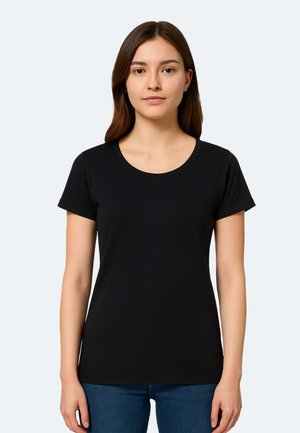 Jeune femme aux cheveux bruns et lisses portant un T-shirt noir uni à manches courtes et un jean bleu, debout devant un fond clair.