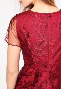 Bordeauxfarbene Kleid mit durchsichtigen, bestickten Ärmeln, die mit strukturierten, blätterartigen Mustern verziert sind. Reißverschluss hinten und taillierte Passform mit Falten_detail.