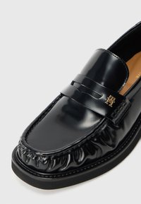 Mocassim de couro preto brilhante com bico arredondado, detalhes cosidos e um pequeno emblema decorativo dourado na tira.