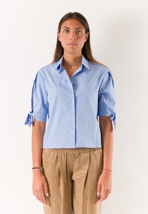 Giovane donna con lunghi capelli scuri che indossa una camicia celeste abbottonata con maniche corte annodate e pantaloni beige larghi su uno sfondo semplice.