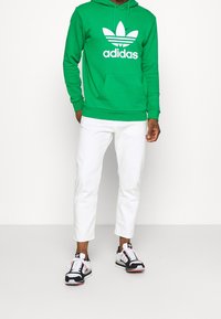 Zelená mikina s bílým logem Adidas, bílé kalhoty a černé/červené tenisky. Mikina má přední kapsu a kapuci se stahovací šňůrkou.