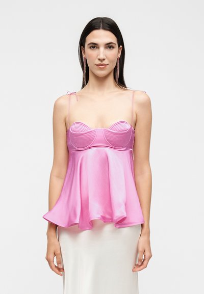 Femme aux longs cheveux foncés portant un caraco en satin rose à fines bretelles et ourlet évasé, assorti à une jupe blanche.