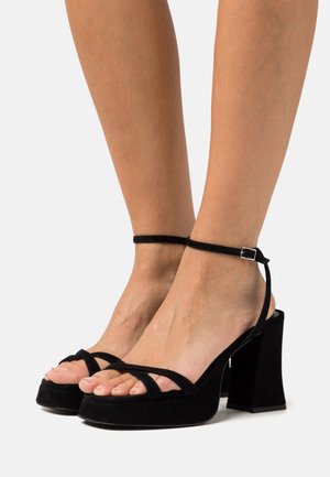Black suede heeled sandals with a block heel, featuring elegant cross-straps and an adjustable ankle strap with a silver buckle detail.