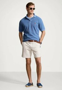 Vyras vilkintis mėlyną polo marškinėlį, šviesias trumpas kelnes, nertas rudą diržą, mėlynus atviro tipo batus ir saulės akinius, stovintis su rankomis kišenėse.