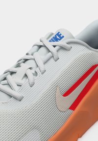 Λευκό αθλητικό παπούτσι Nike με μπλε λογότυπο στη γλώσσα, μπεζ σούρπας, κόκκινη ρίγα διακοσμητική, λευκά κορδόνια και πορτοκαλί σόλα σε κοντινή λήψη.