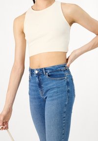 Personne portant un crop top blanc sans manches et un jean bleu taille haute, tenant une sangle beige d'une main et l'autre main sur la hanche.