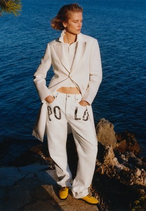 Fehér blézert, „POLO” feliratú fehér nadrágot és sárga cipőt viselő nő áll a sziklás parton, kék víz mellett, oldalra nézve.
