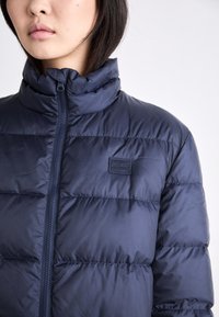 Tumedavine puffy jakk, millel on kõrge krae, lukuga esikülg ja õmmeldud sektsioonid. Rinnal on tekstuuritud logo patch.