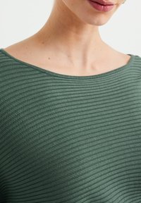 Grön ribbad tyg med bred u-ringning, som visar en texturerad yta och slät finish, med fokus på bröstområdet.