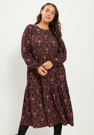Mujer con cabello largo y castaño vistiendo un vestido midi de manga larga con un patrón en rojo oscuro y negro, de pie con las manos entrelazadas delante.