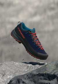 Kék és fekete túracipő, textúrált felülettel, élénk narancssárga fűzőkkel kiemelve. Könnyű kialakítás, kültéri tevékenységekhez megfelelő, sziklás felület felett látható.