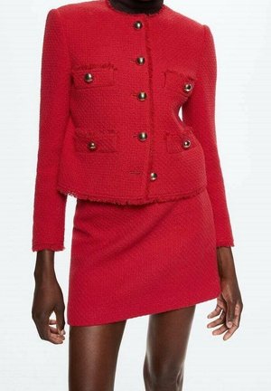 Femme portant une veste rouge texturée sur mesure avec des boutons dorés, assortie à une mini-jupe rouge, sur un fond uni.