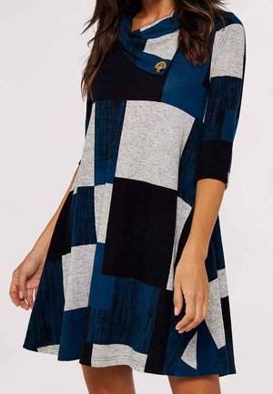 Femme portant une robe mi-longue à manches trois-quarts, avec un motif patchwork composé de blocs bleus, noirs et gris, et un col bénitier boutonné.