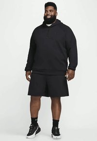 Lächelnder Mann in schwarzem Hoodie, schwarzen Shorts, schwarzen Socken und schwarz-weißen Sneakern, der vor einem schlichten Hintergrund steht.