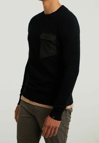 Černý pletený svetr s texturovaným povrchem a přední náprsní kapsou. Obsahuje žebrované manžety a spodní lem, nošený přes béžovou košili.