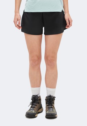 Asmuo, vilkintis juodus sportinius šortus, baltas kojines ir rudus bei juodus žygių batus, stovintis kojas šiek tiek išskėtęs.