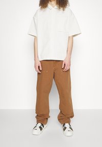 Světlý béžový tričko s krátkým rukávem, knoflíkovým límcem a kapsou, spárované s hnědými volně střiženými kalhotami a bílými teniskami s černými akcenty.