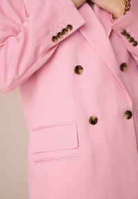 Dvouřadový růžový blazer z lehkého materiálu, s knoflíky ve zlatém odstínu, bočními kapsami a hladkou texturou.