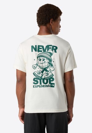 Mand med en kridhvid T-shirt med grøn grafik af en gangende jordklode i vinterhat og støvler, teksten "NEVER STOP EXPLORING" på ryggen.