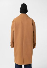 Persoon met een lange camelkleurige jas en een zwarte muts, met de rug naar de kijker, tegen een effen lichtgrijze achtergrond.