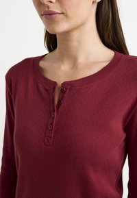 Blusa burdeos de manga larga hecha de tejido texturizado, con un cuello redondo, placket de botones y pequeños patrones de diamante en toda su superficie.