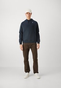 Felpa navy con tasca a marsupio, logo bianco sul petto, abbinata a chinos marroni e sneakers bianche; il modello indossa un cappellino chiaro.
