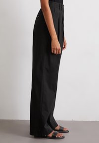 Zwarte wijde broek van een katoenmix, met voorplooien en zakken aan de zijkant. Model draagt zwarte platte sandalen.