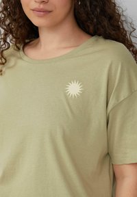Olivgrünes Baumwoll-T-Shirt mit einem runden Halsausschnitt und kurzen Ärmeln; mit einem hellen Sonnenmotiv auf der linken Brust.