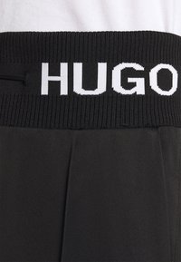 Černé sportovní šortky s žebrovaným pasem, na kterém je bílé logo "HUGO". Tkanina vypadá hladce a má volnější střih.