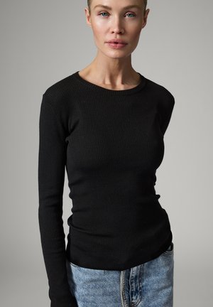 Femme à la peau claire et aux yeux bleus portant un haut noir côtelé ajusté à manches longues et un jean bleu clair sur un fond uni.