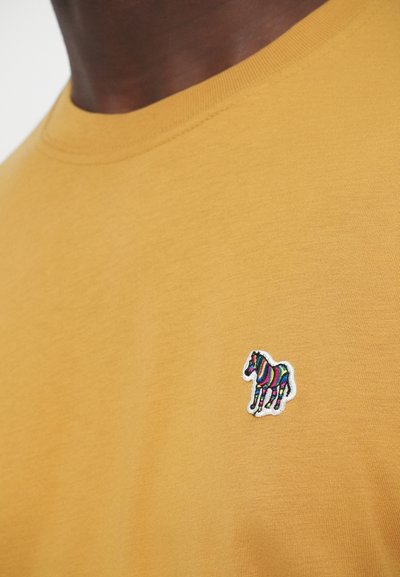 Žlté bavlnené tričko s okrúhlym výstrihom a malým vyšívaným viacfarebným logom zebry na ľavej strane hrudníka. Mäkká textúra.