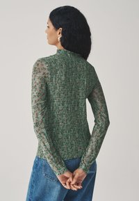 Green long-sleeve top with sheer floral sleeves, textured bodice, and gathered back design. Worn with high-waisted denim jeans.