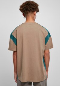 Beige Kurzarm-T-Shirt mit türkisfarbenen Akzenten, aus weichem Baumwollmaterial. Es hat eine lockere Passform und kontrastierende Farbakzente an den Schultern.