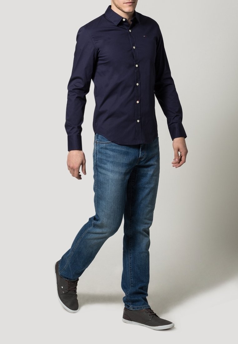 Navy blue long-sleeve shirt with a button-down collar, light buttons, paired with faded blue jeans and gray casual shoes.