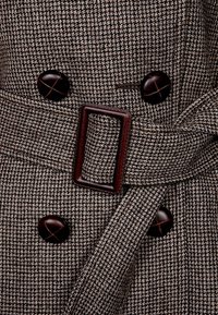 Płaszcz w brązowo-szary wzór houndstooth z szerokim paskiem materiałowym, dużymi okrągłymi guzikami i kwadratową klamrą paska. Teksturowana powierzchnia materiału.