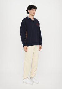 Sweatshirt azul-marinho com decote em V, detalhes branco e castanho, e bordado. Combinado com calças de moletom na cor creme e ténis brancos.