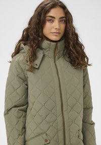 Chaqueta acolchada verde oliva con cuello alto, cierre de cremallera, bolsillos y botones a presión. Presenta un patrón texturizado y capucha.