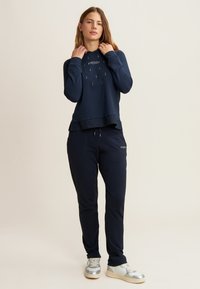 Felpa blu navy con cappuccio, dettaglio del logo e design a strati abbinata a pantaloni jogger coordinati. Il tessuto appare morbido e liscio.