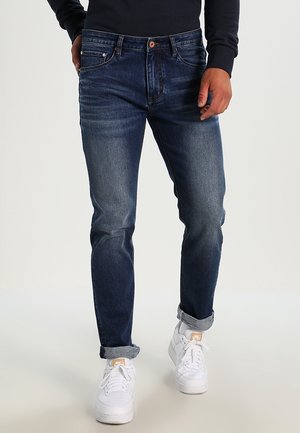 Mężczyzna noszący ciemnoniebieskie dopasowane jeansy z podwiniętymi nogawkami, w połączeniu z białymi sneakersami i czarnym długim rękawem, stojący na jednolitym tle.